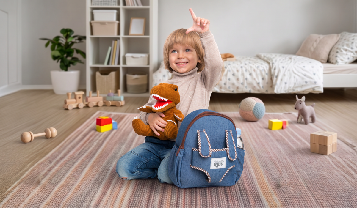 Toddler hugs a corduroy plush T-Rex next to a Naturally KIDS small backpack with a water bottle in the side pocket