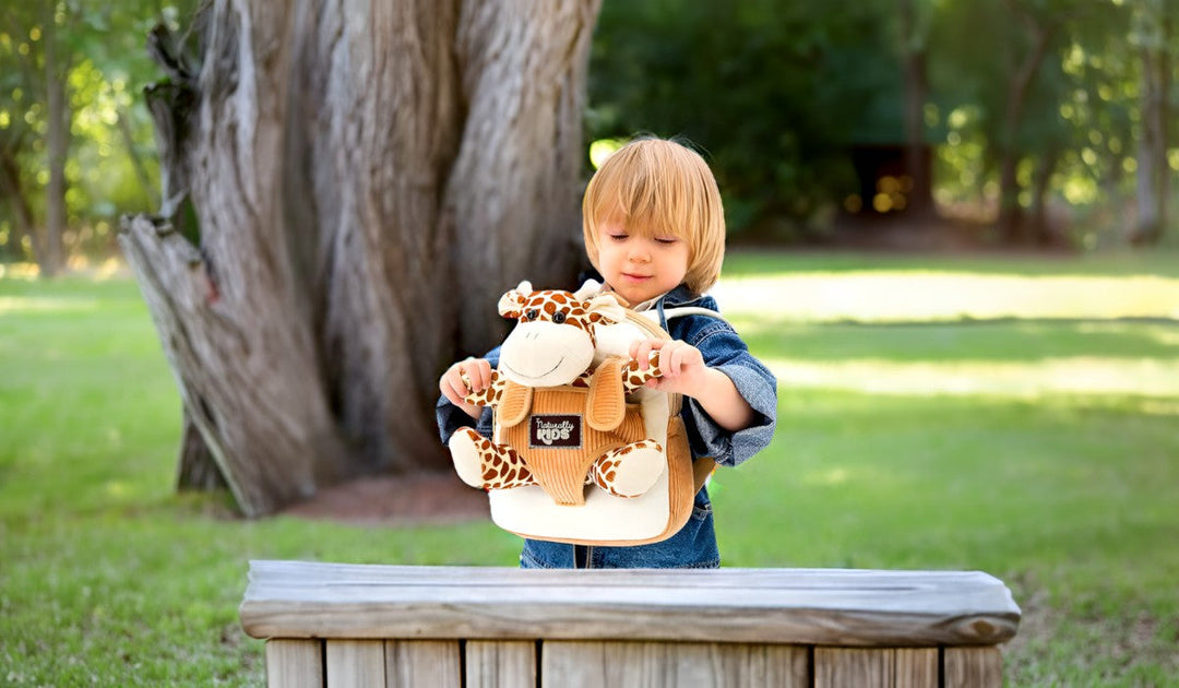 Toddler holds a Naturally KIDS small backpack with a plush giraffe in front, outdoors by a tree