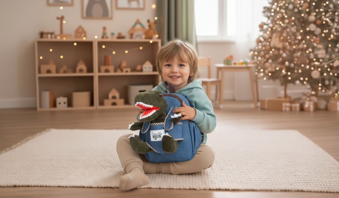 A toddler sits on a rug by a Christmas tree, hugging a Naturally KIDS blue backpack with a green plush T-Rex