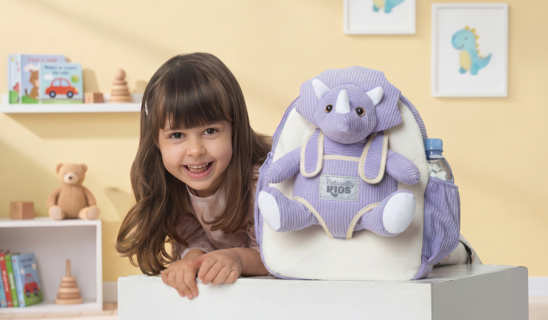 A smiling child leans on a table next to a Naturally KIDS backpack with pockets and a plush purple triceratops